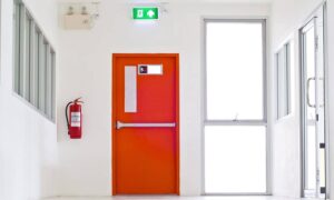 A hallway with a red fire door, a fire extinguisher and emergency lighting